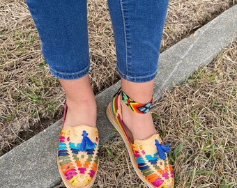 May include: A pair of colorful woven leather sandals with blue tassels. The sandals have a strap that goes around the ankle and are worn with blue jeans.