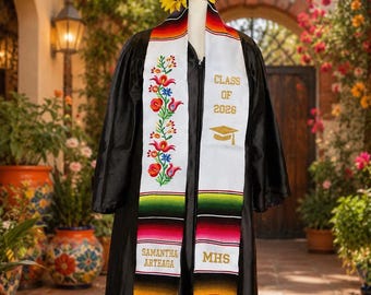 May include: A black graduation gown with a white embroidered stole featuring colorful floral patterns, the text "CLASS OF 2026", "SAMANTHA ARTEAGA", and "MHS", a gold graduation cap icon, and a multi-colored striped border with fringe, topped with sunflowers.