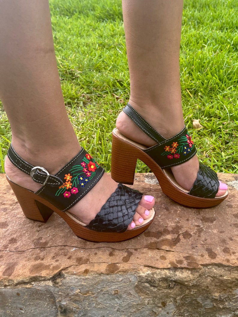 Puede incluir: Sandalias de plataforma de cuero negro con correas tejidas y tac&oacute;n de madera. Las sandalias presentan bordados florales coloridos en las correas y cierre de hebilla. El tac&oacute;n mide aproximadamente 7,6 cm.