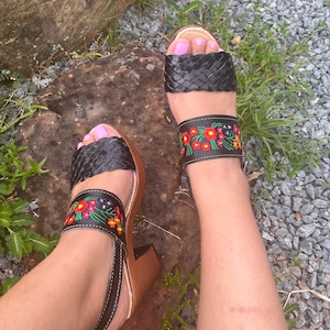 Puede incluir: Sandalias de cuero negro con correas trenzadas en los dedos y correas de tobillo bordadas con flores. Las sandalias tienen un tac&oacute;n de madera y una correa en el tobillo. Llevadas sobre una superficie de piedra.