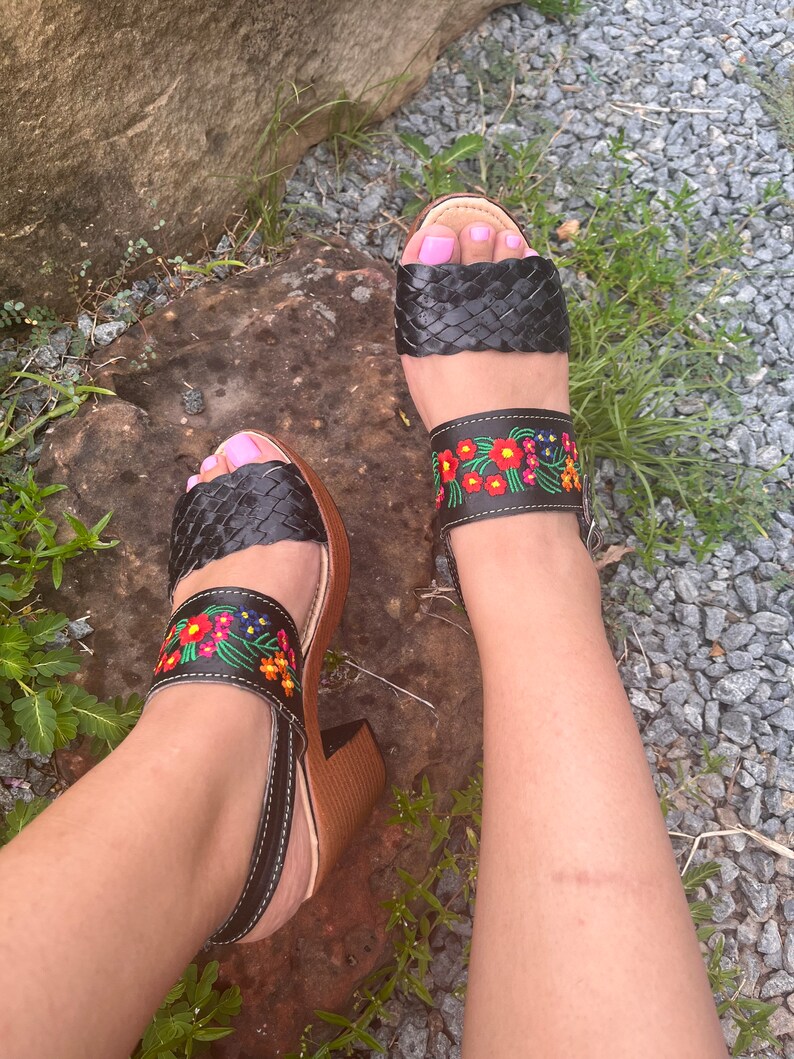 Puede incluir: Sandalias de cuero negro con correas trenzadas en los dedos y correas de tobillo bordadas con flores. Las sandalias tienen un tac&oacute;n de madera y una correa en el tobillo. Llevadas sobre una superficie de piedra.