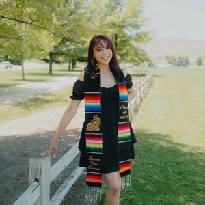 Puede incluir: Una mujer con un vestido negro y una banda de graduación colorida. La banda tiene el texto "Class of 2023" y "Azucena Ruiz Gracias" bordado.