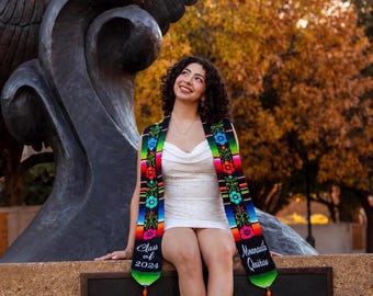 Può includere: Una stola di laurea con base nera e vivaci ricami floreali multicolori di ispirazione messicana e motivi a righe. Il lato sinistro riporta il testo 'Class of 2024' e il lato destro il nome 'Miranda Quiroz' con nappe di colore arancione.