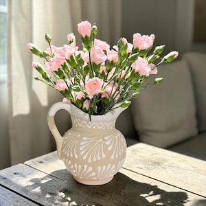 May include: A ceramic pitcher with a cream-colored base and a white handle, featuring a repeating white fan-shaped pattern around the body and a scalloped line near the rim, filled with a bouquet of light pink carnations and green buds.
