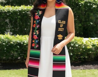 May include: A black graduation stole featuring vibrant floral embroidery on one side and the gold text "CLASS OF 2027" with a matching graduation cap icon on the other. The stole includes colorful horizontal stripes in green, pink, and orange.