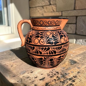 Puede incluir: Una jarra de cerámica color terracota con cuerpo redondeado, asa curva y pico vertedor. La superficie está decorada con intrincados patrones negros pintados a mano, que incluyen volutas, motivos de hojas y bandas horizontales.