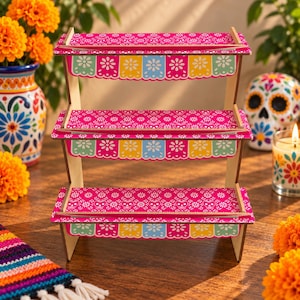 May include: A three-tiered wooden display stand featuring a vibrant pink surface with a white skull and floral pattern. The front edges of each shelf are decorated with a colorful scalloped banner design in shades of yellow, blue, and green.