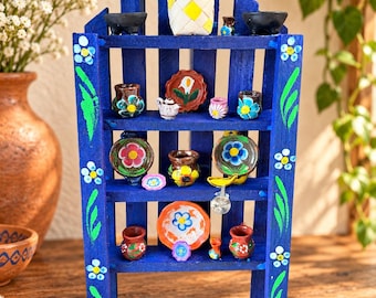 Pode incluir: Uma prateleira de exposição em madeira azul com topo pontiagudo, pintada à mão com folhas verdes e flores brancas. As quatro prateleiras contêm um sortido de objetos de cerâmica em miniatura, incluindo pequenas taças, chávenas, pratos e um cesto.