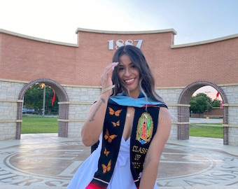 May include: A woman wearing a white dress and a graduation sash with butterflies and the text "Class of 2023". She is smiling and holding her graduation cap in her hand. The background is a brick building with an archway.