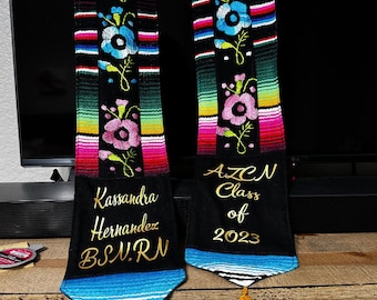 May include: Two graduation sashes with colorful stripes and embroidered flowers. The sash on the left has the name "Kassandra Hernandez" and the text "BSN, RN" embroidered in gold. The sash on the right has the text "AZCN Class of 2023" embroidered in gold.