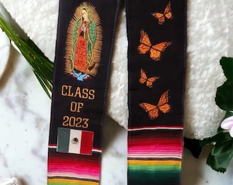 May include: Black graduation stole with a colorful Mexican serape pattern at the bottom. The stole features an image of the Virgin Mary, the text "CLASS OF 2023", and the Mexican flag. The stole also has a row of orange butterfly designs.