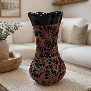 Puede incluir: Un jarrón de terracota con un borde festoneado negro brillante y un patrón floral pintado en negro que cubre el cuerpo. El jarrón presenta un cuello estrecho que se ensancha hacia arriba y una base redondeada, mostrando el contraste mate.
