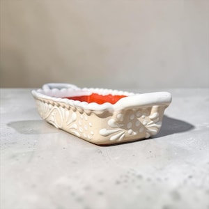 May include: A rectangular ceramic baking dish with a light beige exterior featuring raised white floral and dot patterns. The dish has two white handles on the ends and a smooth, vibrant orange interior finish with a scalloped white rim edge.
