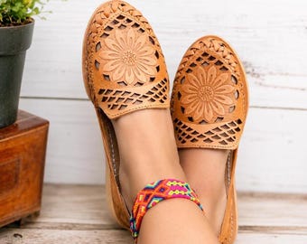 Puede incluir: Un par de zapatos planos huarache de cuero color canela con un diseño de rejilla trenzada y un patrón de girasol en relieve en la parte superior. Los zapatos se llevan con una tobillera de tela tejida multicolor con formas geométricas.