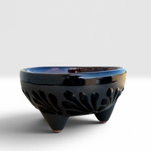 May include: A small, three-legged ceramic bowl with a dark, matte black exterior featuring a carved, repeating leaf-like pattern. The interior of the bowl has a smooth, glossy dark finish, and the three short feet reveal a natural terracotta base.
