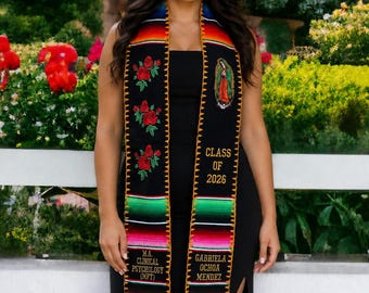 May include: A black graduation stole featuring colorful serape-style stripes at the ends, red rose embroidery, and an image of the Virgin of Guadalupe. Text includes "CLASS OF 2026", "M.A. CLINICAL PSYCHOLOGY (MFT)", and "GABRIELA OCHOA MENDEZ" in gold.