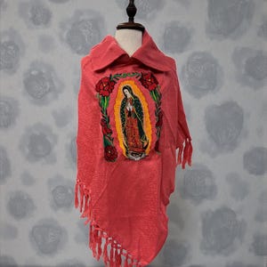 May include: A coral-colored poncho with a detailed embroidered image of the Virgin Mary, framed by red roses and green leaves. The poncho has a textured weave, a collar, and fringe along the bottom edge. The garment is displayed on a mannequin.