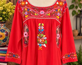 Op de afbeelding: Een felrode blouse met lange mouwen, een wijde ronde halslijn en ingewikkeld bloemenborduurwerk. Het borduurwerk toont levendige, veelkleurige bloemen en bladeren op de borst, in het midden en langs de manchetten van de mouwen.