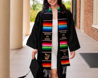 May include: A black graduation gown worn over a white dress, featuring a colorful serape stole with horizontal stripes. The stole displays the text 'SAM HOUSTON STATE UNIVERSITY', 'CLASS OF 2026', and 'PARA MI ABUELO', along with gold butterfly embroidery.