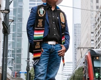 May include: A white cowboy hat, a black button-down shirt, a blue blazer, and blue jeans. A graduation stole with colorful stripes, the text "GRACIAS A MIS PADRES POR TODO", "JOSE RAMOS", and "CLASS OF" is worn over the blazer, featuring religious imagery.