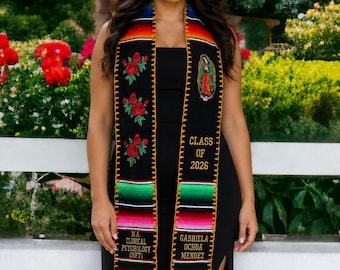May include: A black graduation stole featuring colorful serape-style stripes at the ends, red rose embroidery, and an image of the Virgin of Guadalupe. Text includes "CLASS OF 2026", "M.A. CLINICAL PSYCHOLOGY (MFT)", and "GABRIELA OCHOA MENDEZ" in gold.