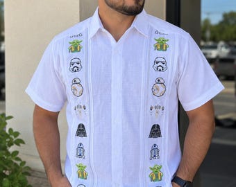 Pode incluir: Uma camisa branca de manga curta com botões, apresentando dois painéis verticais de personagens de Star Wars bordados em pixel art, incluindo Yoda, soldados imperiais, BB-8, Darth Vader e R2-D2, dispostos num padrão repetitivo na frente.