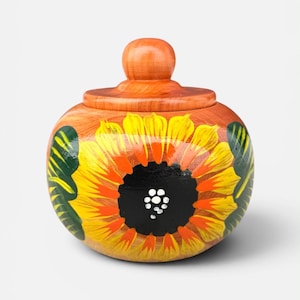 Peut inclure: Un pot en bois avec un couvercle, peint à la main avec un motif de tournesol. Le pot est rond avec une finition marron clair. Le tournesol présente des couleurs jaune, orange et noir. Des feuilles vertes sont peintes sur les côtés.