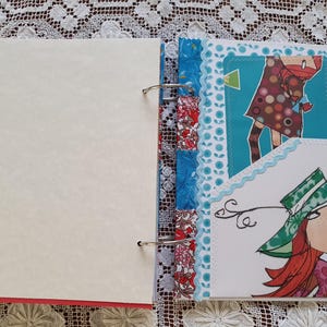 May include: A scrapbook with a white cover and a colorful patterned spine. The scrapbook is open to a page with a blue background and a white card with a drawing of a woman with red hair wearing a green hat. The card is decorated with white lace.