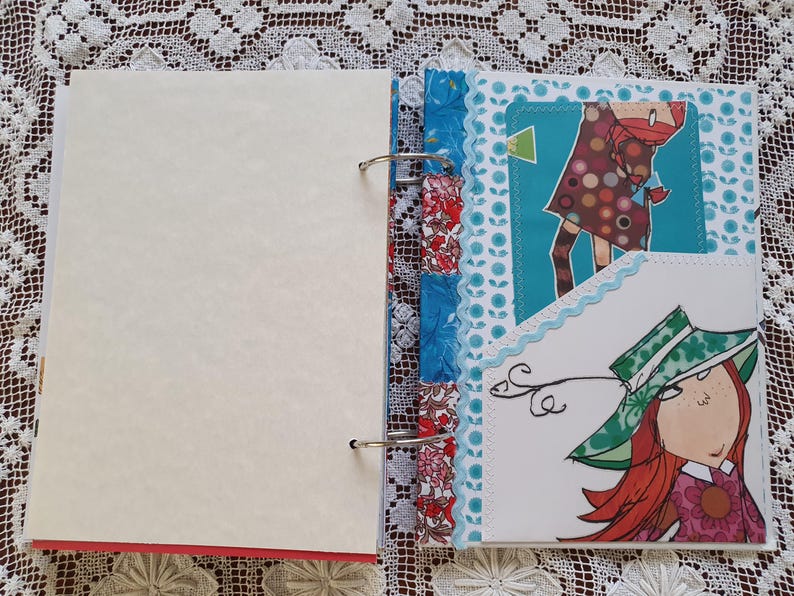 May include: A scrapbook with a white cover and a colorful patterned spine. The scrapbook is open to a page with a blue background and a white card with a drawing of a woman with red hair wearing a green hat. The card is decorated with white lace.