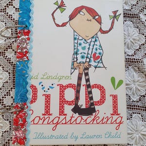 May include: A white book with a red and blue patterned cover. The cover features a cartoon illustration of a girl with red hair and a blue dress. The title of the book is "Pippi Longstocking" and the author is "Astrid Lindgren". The book is illustrated by Lauren Child.