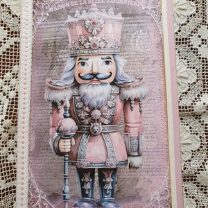 Puede incluir: Una figura de cascanueces rosa y blanca con una corona, bigote y barba. La figura sostiene un bastón con una parte superior redonda. El fondo es un diseño de estilo vintage con texto en francés.