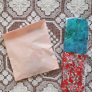 May include: A pink paper bag with white polka dots next to a stack of fabric scraps. The top scrap is a teal blue with white stripes, the middle scrap is a floral print with red, pink, and white flowers, and the bottom scrap is a solid red fabric.