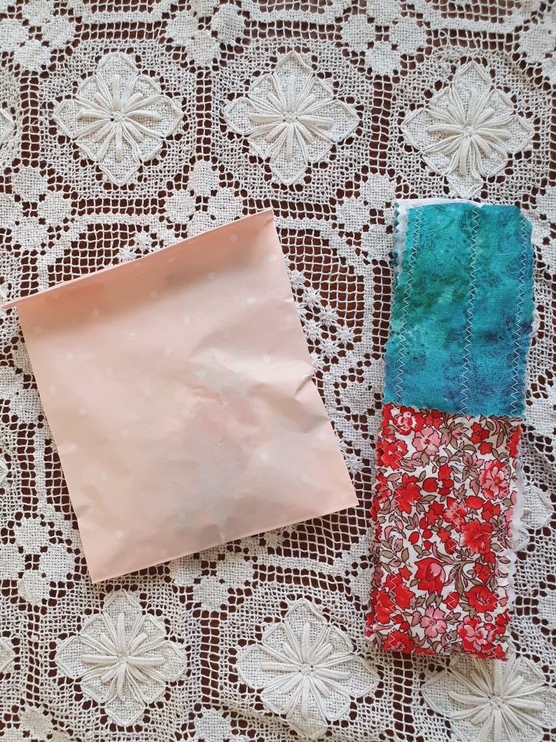 May include: A pink paper bag with white polka dots next to a stack of fabric scraps. The top scrap is a teal blue with white stripes, the middle scrap is a floral print with red, pink, and white flowers, and the bottom scrap is a solid red fabric.