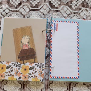 May include: A brown and beige scrapbook with a floral pattern on the bottom. The left page features a cartoon illustration of a girl sitting at a desk. The right page has a blank white space with a blue and red airmail border.