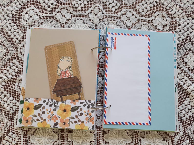May include: A brown and beige scrapbook with a floral pattern on the bottom. The left page features a cartoon illustration of a girl sitting at a desk. The right page has a blank white space with a blue and red airmail border.