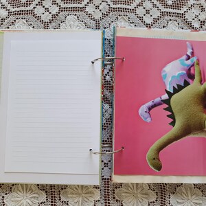 May include: An open binder with a white lined page on the left and a pink page with two dinosaur plushies on the right. The dinosaurs are green and purple with a pink background.