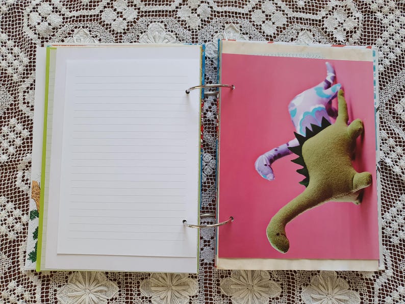 May include: An open binder with a white lined page on the left and a pink page with two dinosaur plushies on the right. The dinosaurs are green and purple with a pink background.