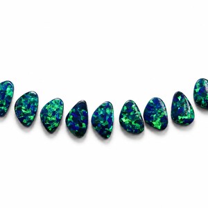 Puede incluir: Una colección de once cabujones en forma de lágrima dispuestos en un arco suave. Cada piedra presenta una base azul oscuro con destellos iridiscentes vibrantes de verde brillante y azul eléctrico en toda la superficie pulida y lisa.