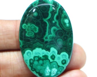 May include: Oval-shaped malachite gemstone with a polished surface. The stone features a swirling pattern of dark and light green hues, creating a unique, natural design. The gemstone is held in a hand against a white background.