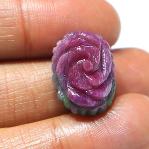Puede incluir: Una gema tallada en forma de rosa. La gema presenta tonos de rosa, morado y verde. El tallado de la rosa es detallado, con pétalos visibles y una superficie texturizada. La gema se sostiene entre los dedos.