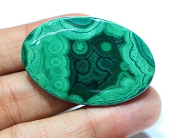 May include: Oval-shaped malachite gemstone with a polished surface. The stone features a vibrant green color with intricate swirling patterns in varying shades of green and dark green. The gemstone is held in a hand against a white background.