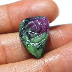 Puede incluir: Una gema tallada con un diseño de rosa. La gema, con forma de corazón, presenta una paleta de colores púrpura oscuro, verde y negro. La talla muestra los pétalos y las hojas de la rosa.