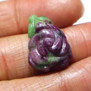 May include: A carved gemstone in shades of deep magenta and green. The stone has a swirling, organic design, with a rounded shape. The gemstone is held in a person's hand, and the background is a neutral colour.