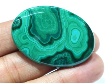 May include: Oval-shaped malachite gemstone with a polished surface. The stone displays a vibrant green color with swirling patterns of lighter and darker shades, creating a natural, organic design. The gemstone is held in a hand.