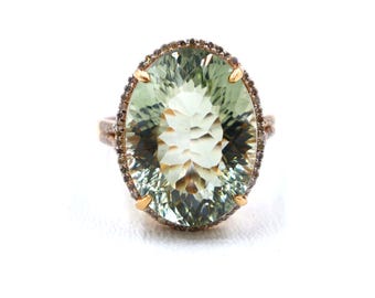 Puede incluir: Anillo ovalado con una gran gema verde claro. La gema está rodeada por un halo de piedras oscuras más pequeñas, engastadas en una banda dorada. El anillo se muestra sobre un fondo blanco.