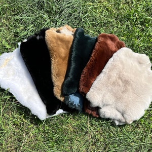 Könnte beinhalten: Sortiment aus sieben Kunstfellstücken in verschiedenen Farben, darunter Weiß, Schwarz, Braun, Beige und Grün. Die Felle sind in einem Halbkreis auf einem Bett aus grünem Gras angeordnet.