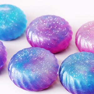 May include: Six round, swirled soaps in shades of blue, purple, and pink, with a glittery, speckled finish. The soaps have a textured, ridged design and are arranged on a white surface.