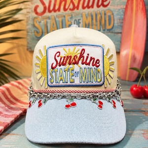 May include: A cream and light blue baseball cap featuring a rectangular embroidered patch that reads Sunshine STATE OF MIND in red and yellow. The cap is accented with a silver-toned chain across the brim, adorned with small red cherry charms.