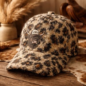 May include: A patterned baseball cap with a brown and tan leopard print design. The cap features an embroidered image of a Highland cow with the word "HOWDY" on a banner above its head. The cap is made of a textured fabric.