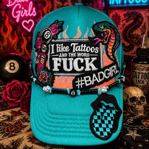 May include: Teal trucker hat with embroidered patches, including a snake, flames, and the text "I like Tattoos and the word FUCK #BADGIRL." The hat features a black beaded band and a checkered tongue patch.